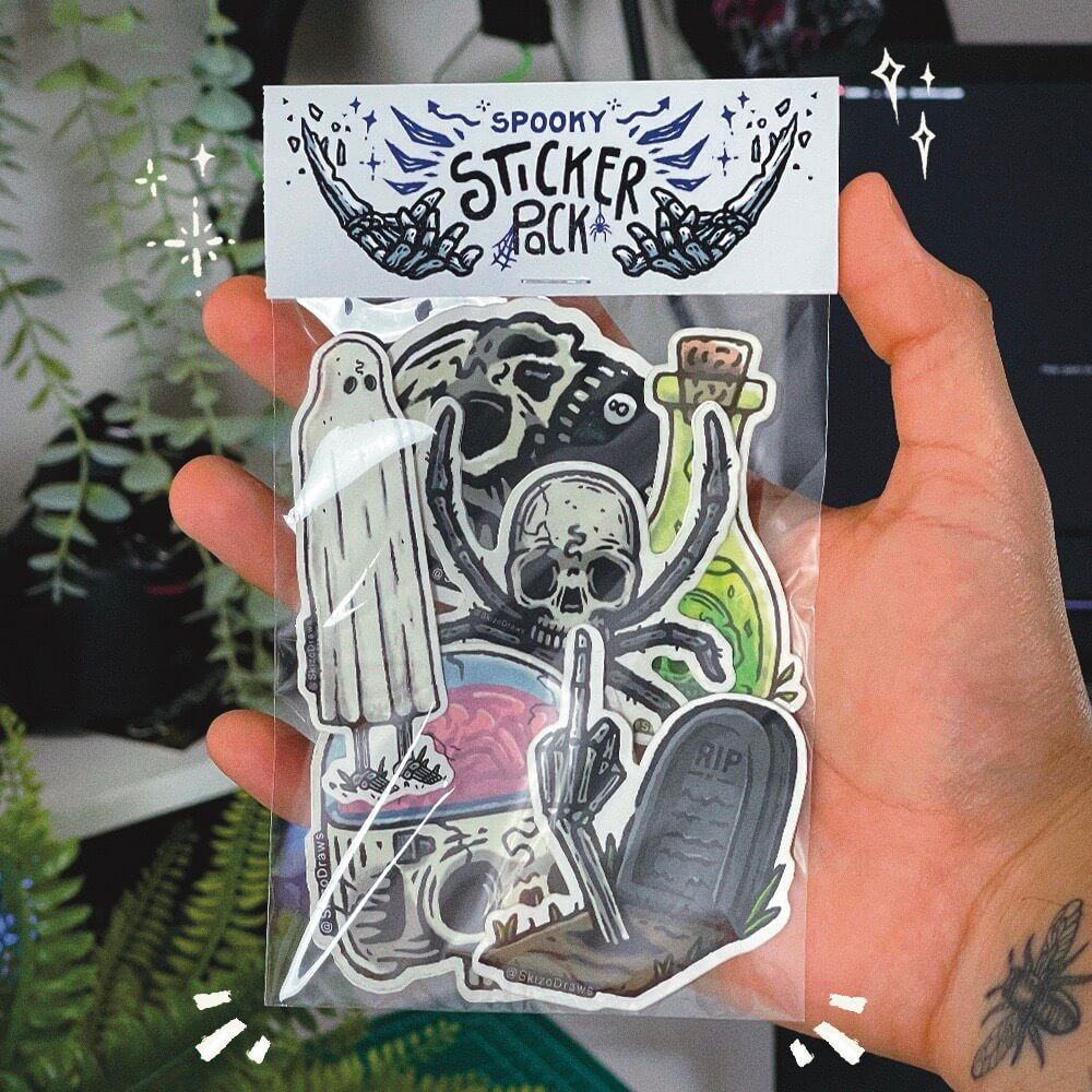 Main tenant un pack de stickers aux dessins sur le thème de l'épouvante, avec une carte d'en-tête indiquant « SPOOKY STICKER PACK », sur fond de plantes.
