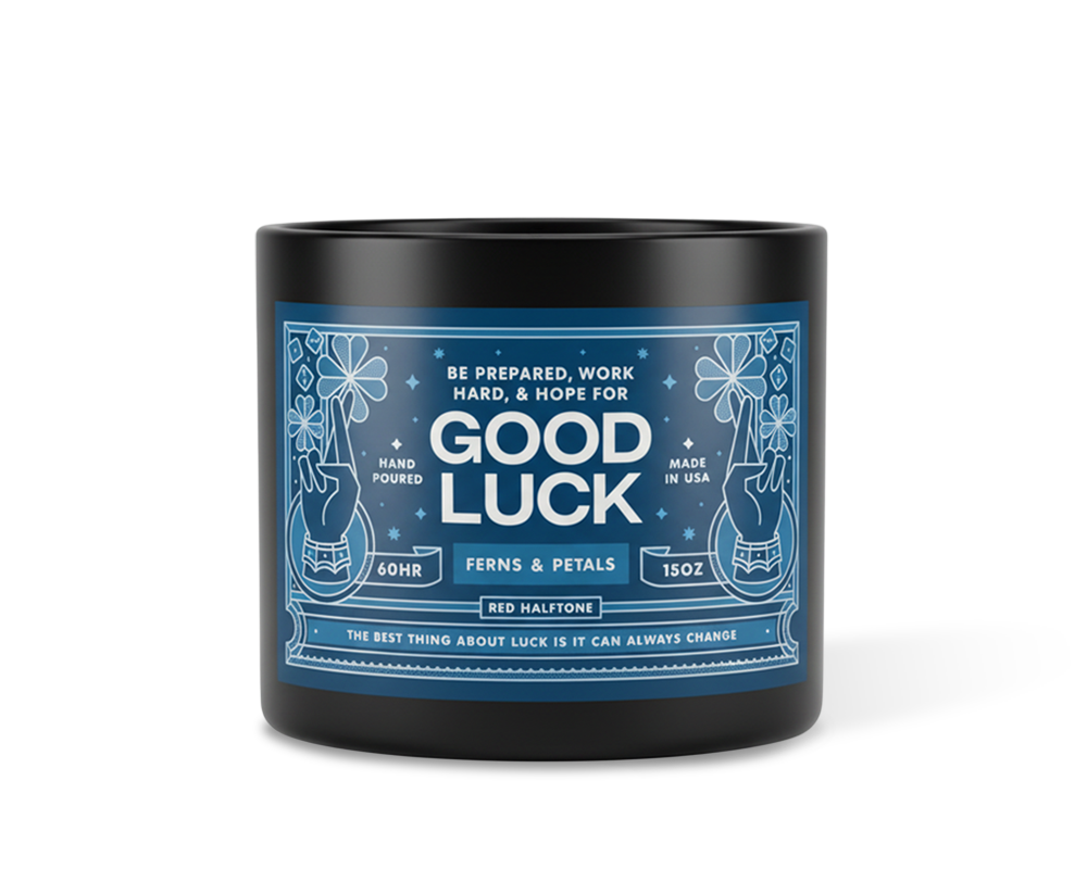 Black candle labeled "Good Luck" with blue and white design featuring ferns and petals. Text includes "Hand Poured" and "Made in USA."