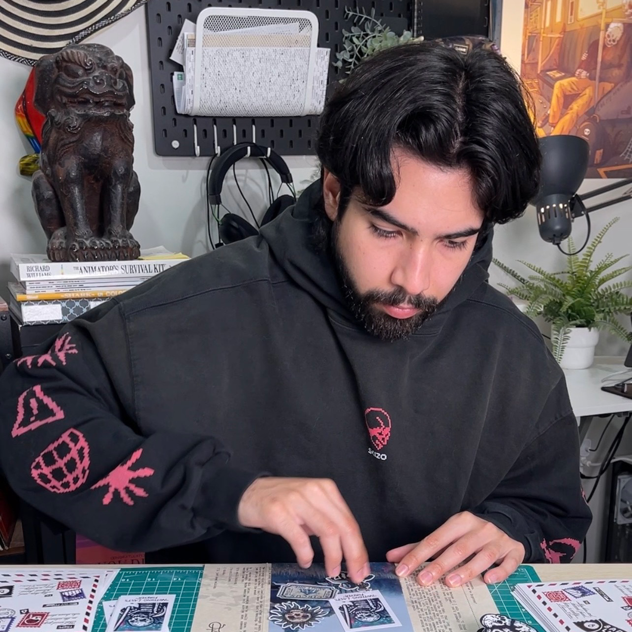 Un homme en sweat à capuche noir, assis dans son atelier, dispose des stickers sur la table devant lui, avec des sculptures et des œuvres d'art en arrière-plan.