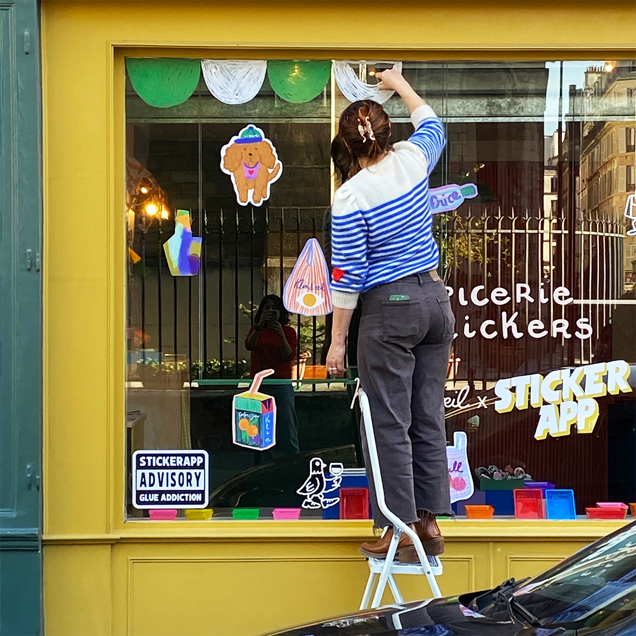 Person in striped sweater on a ladder decorating a window with colorful paper shapes and stickers at a store.