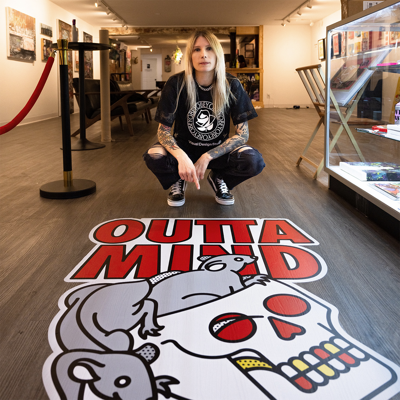 Personne agenouillée à côté d'un sticker coloré "OUTTA MIND" sur le sol d'une galerie, entourée d'œuvres d'art.
