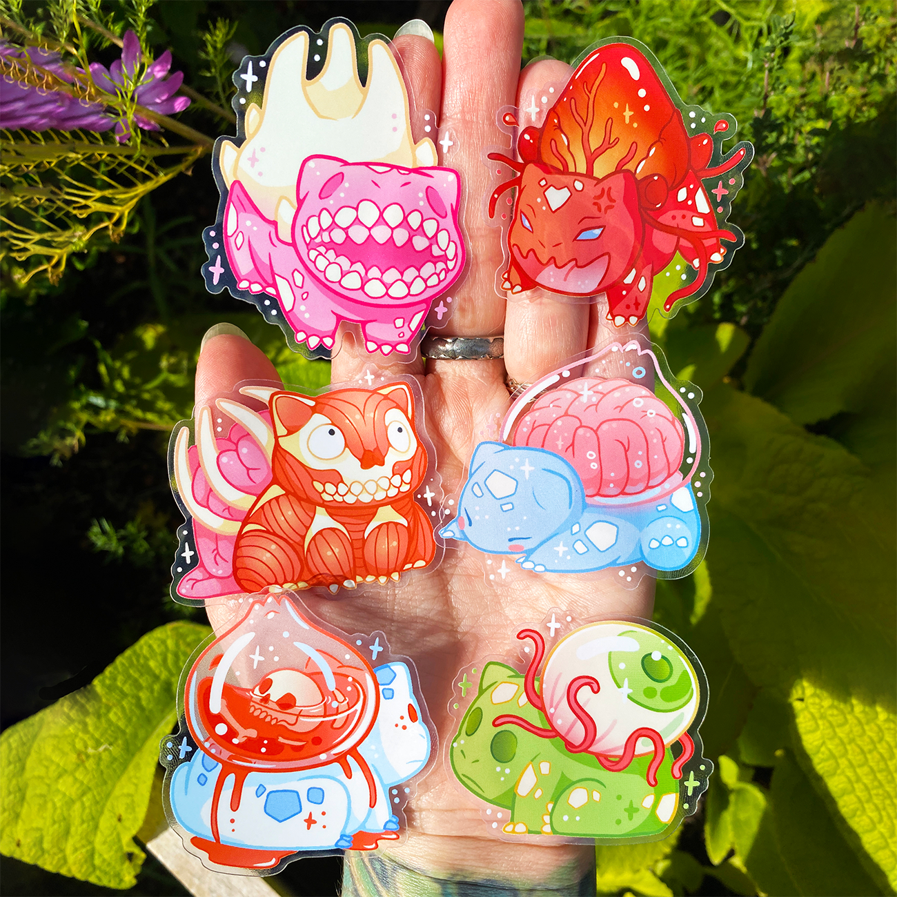 A hand holds six transparent colorful, cartoonish horror bulbasar stickers with various expressions and vibrant designs.