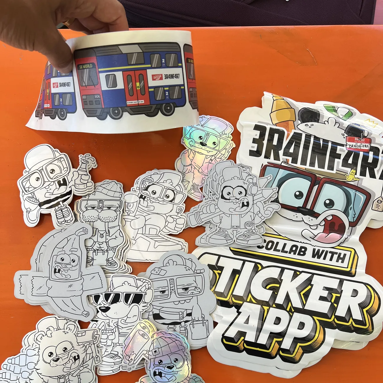 Étiquette du train S8 World Brainfart x StickerApp avec le panneau « In every fart is art », tenue au-dessus de stickers à la forme en noir et blanc