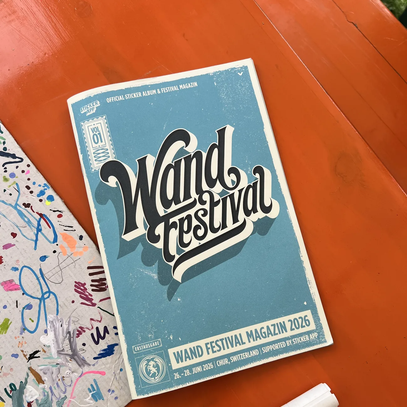 Album de stickers officiel et magazine du Wand Festival 2026, soutenu par StickerApp, à côté d'un marqueur peinture sur une table orange