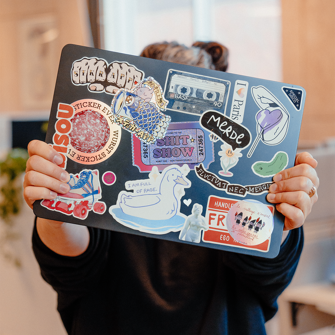 Gros plan d'un ordinateur portable couvert d'un mélange d'autocollants colorés, y compris des personnages illustrés, de la typographie et des designs graphiques, tenu par une personne à l'intérieur.