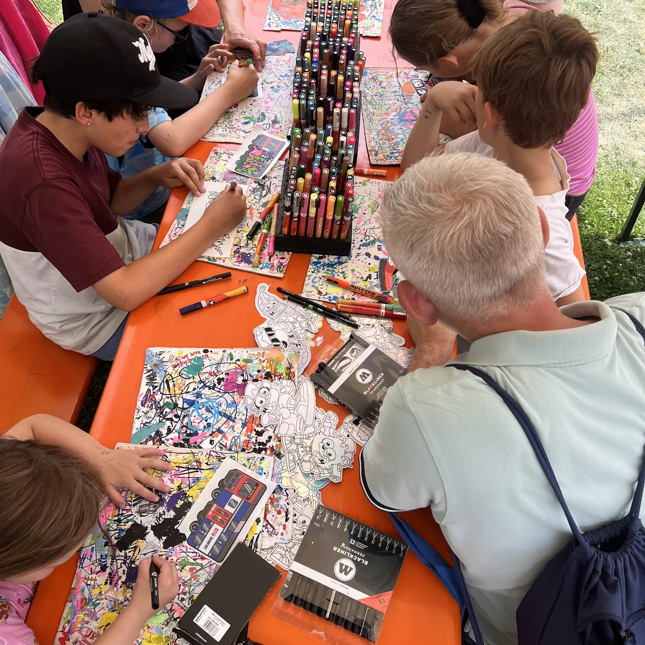 Des enfants colorient des stickers à la forme Brainfart autour d'un présentoir de marqueurs peinture lors d'un atelier de festival en plein air