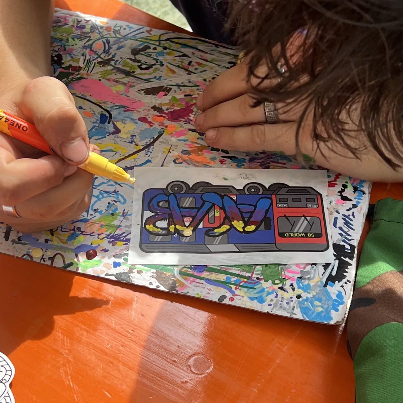 Une main colorie une étiquette du train S8 World au marqueur peinture jaune lors de l'atelier Brainfart x StickerApp