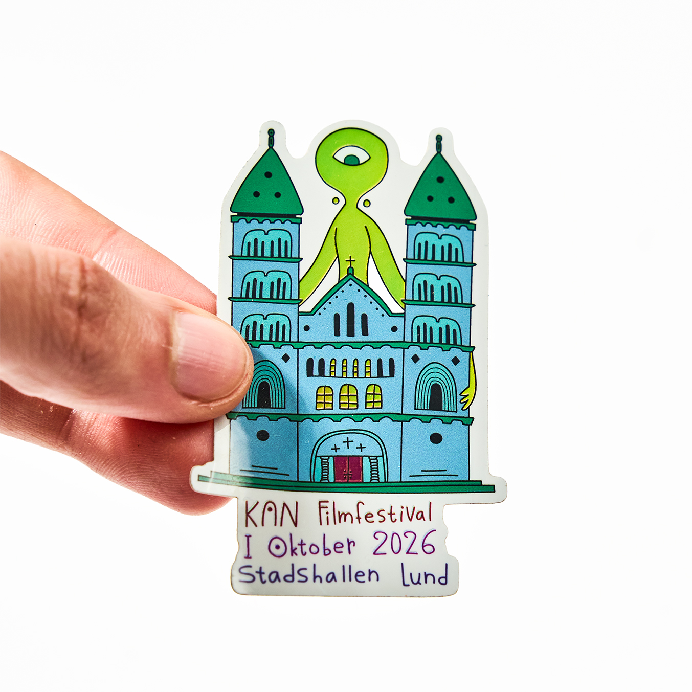 Main tenant un sticker à la forme pour le KAN Filmfestival, octobre 2026 au Stadshallen de Lund, avec une cathédrale et un extraterrestre vert à un œil