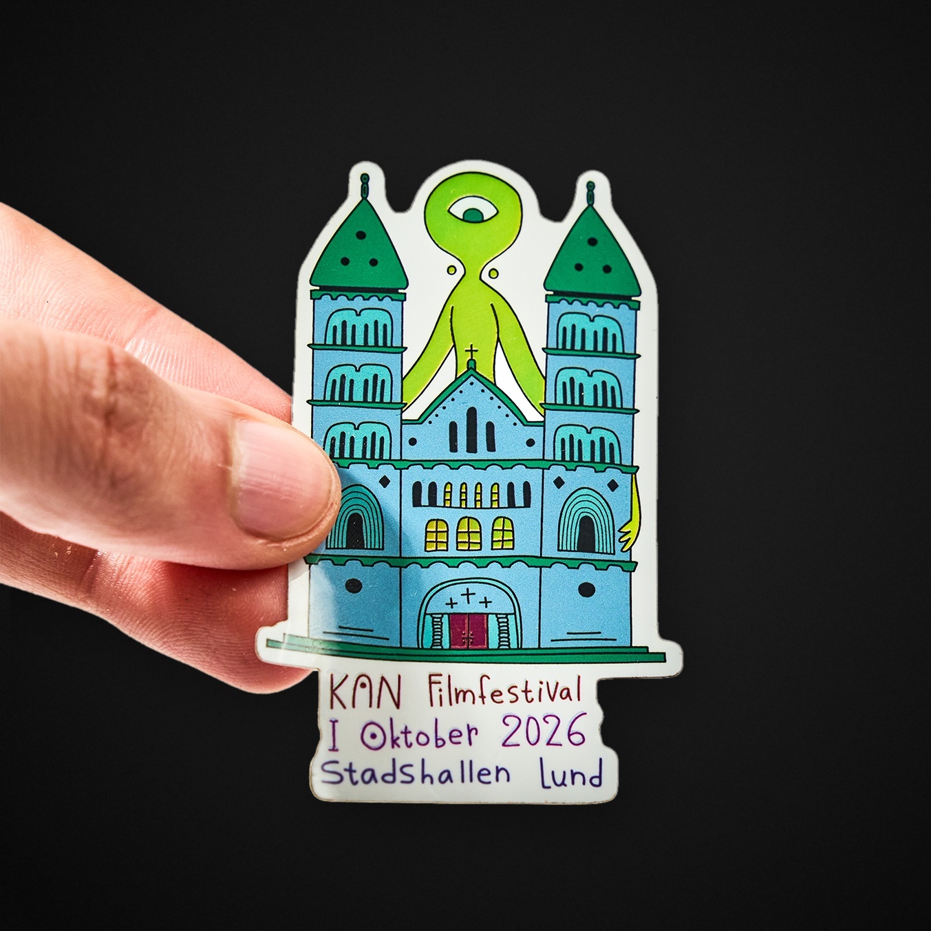 Main tenant un sticker à la forme représentant une cathédrale et une créature verte à un œil, avec le texte « KAN Filmfestival I Oktober 2026 Stadshallen Lund »