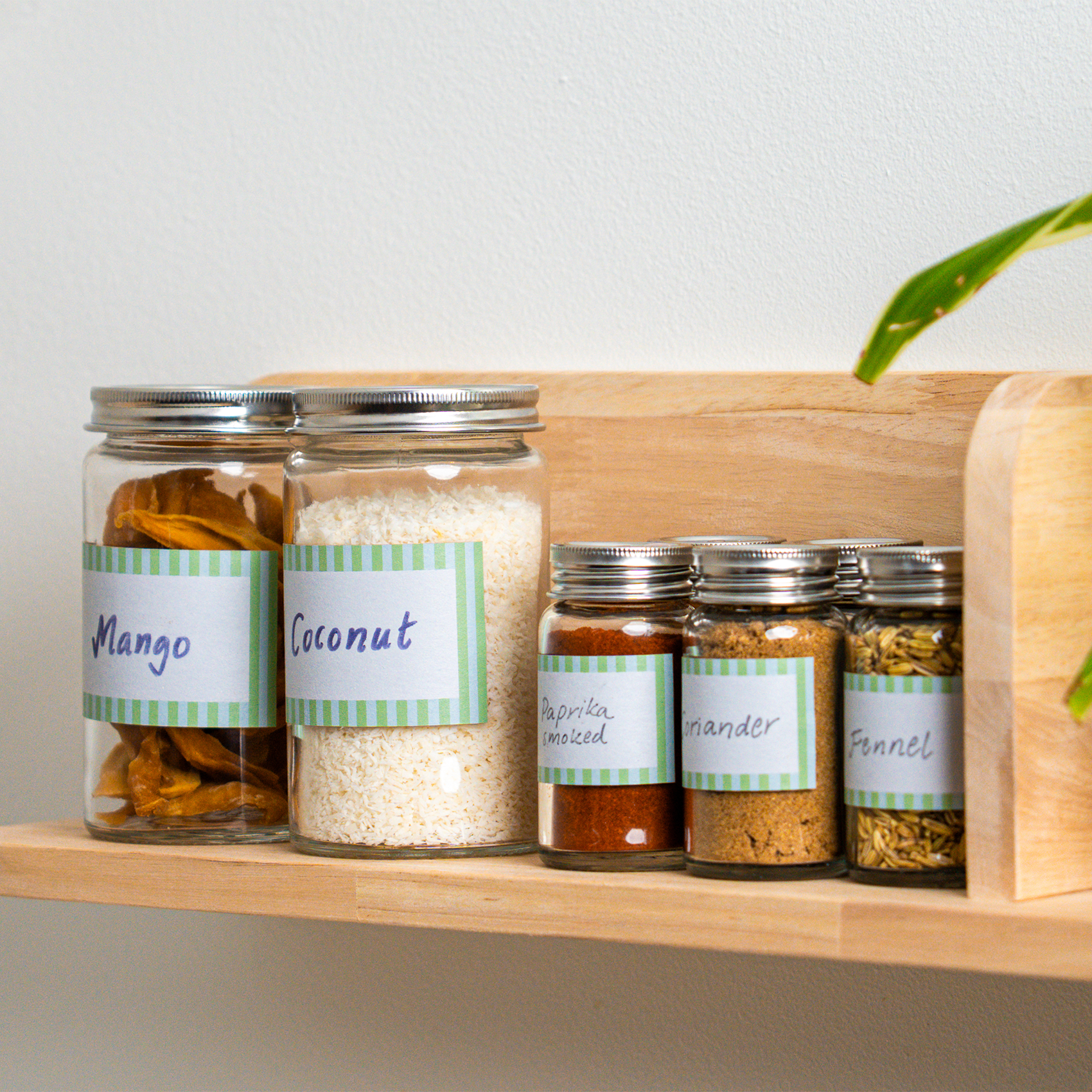 Bocaux en verre avec des étiquettes manuscrites sur une étagère de cuisine en bois. Ils contiennent de la mangue séchée, de la noix de coco et diverses épices, dont du paprika fumé, de la coriandre et des graines de fenouil.
