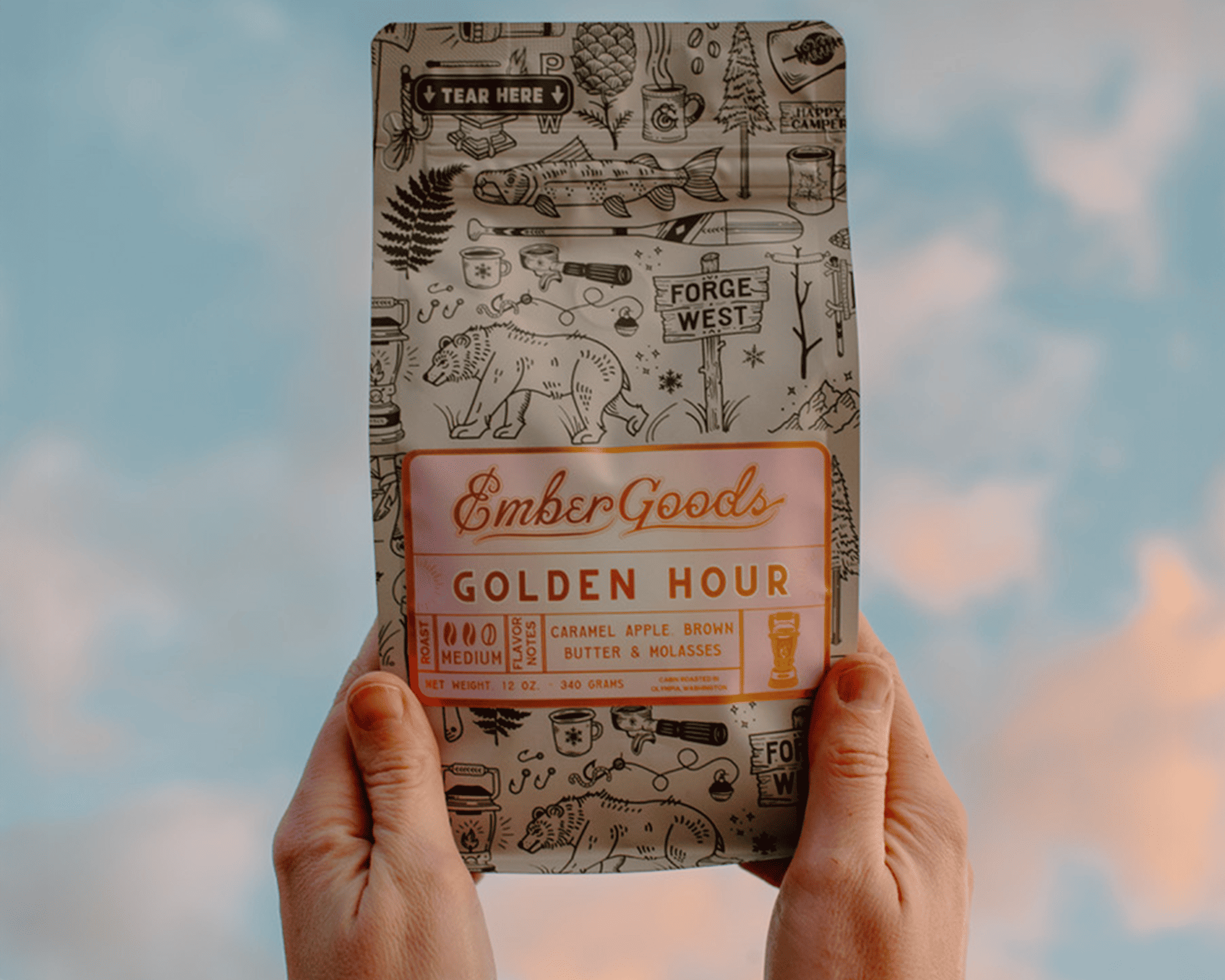 Mains tenant un sachet de café Ember Goods Golden Hour avec une étiquette métallisée sur fond de ciel