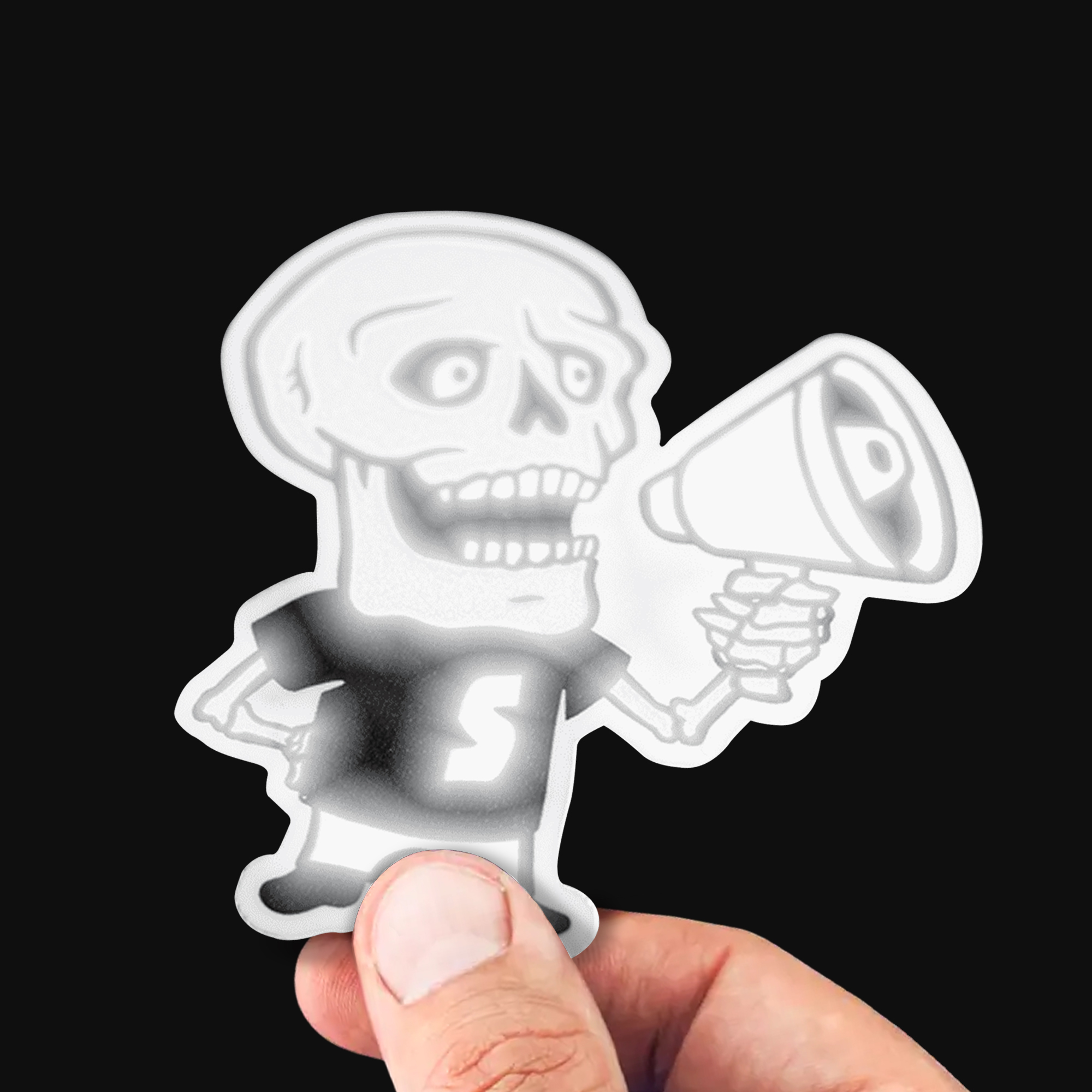 Main tenant un sticker à la forme réfléchissant d'un squelette avec un mégaphone, qui brille en blanc sous une lumière directe